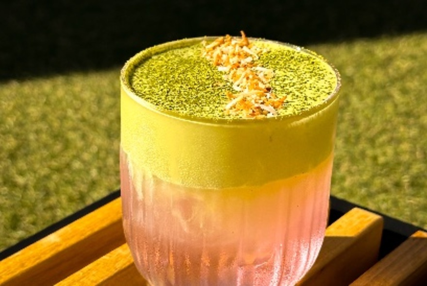 A peachy cocktail with a green foam on a wooden striped board on fake grass. 