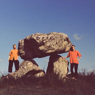 Two people wearing bright orange tops, standing next to balanced standing stones.