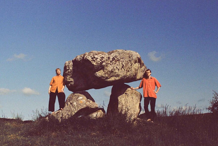 Two people wearing bright orange tops, standing next to balanced standing stones.