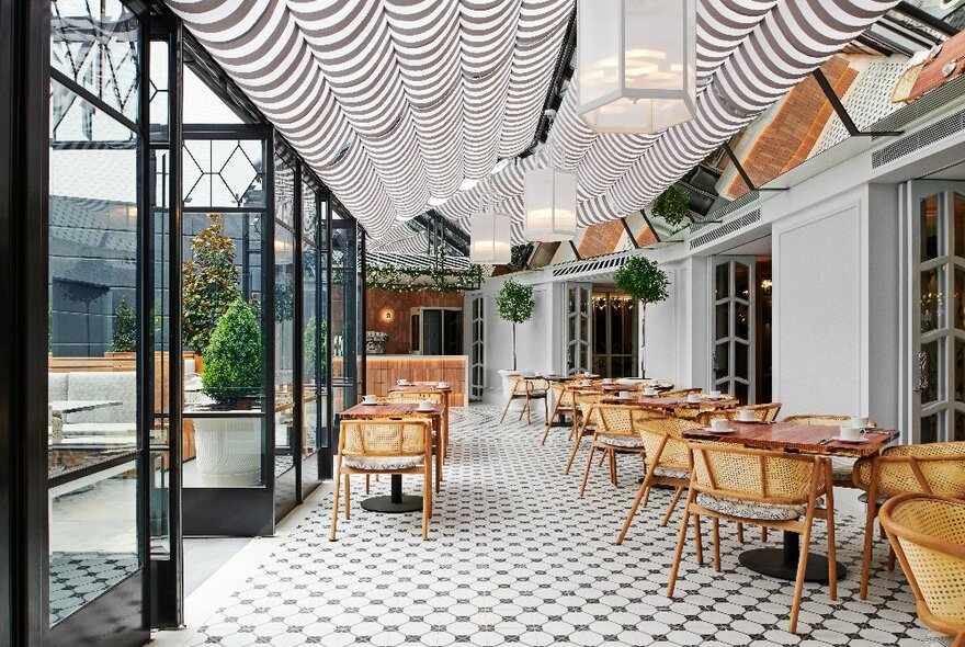 Black and white tiled area, with cane chairs and tables, striped ceiling blinds overhead; wide doors and windows to the left.