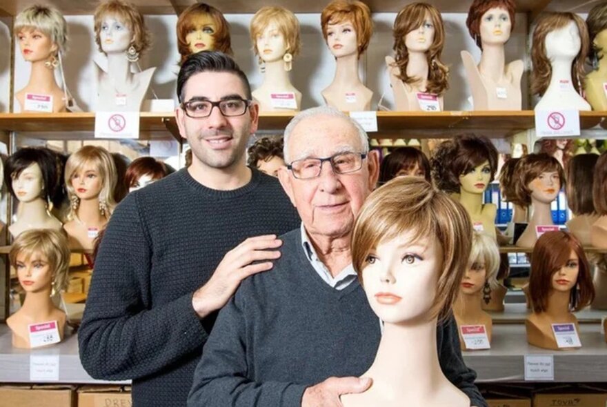 A younger man and an old man holding a bust with a wig on it, the background is shelves of busts with wigs. 