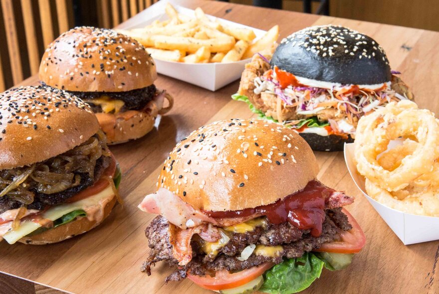Four different types of loaded burgers with a cardboard tray of chips and one of onion rings.