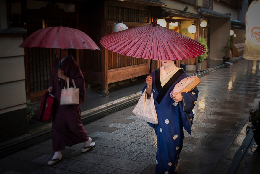 A street scene in Kyoto, Japan, featuring Maiko (apprentice geisha) or Geiko (fully trained geisha) walking along a narrow, paved street in traditional dress, holding dark red parasols over their heads.