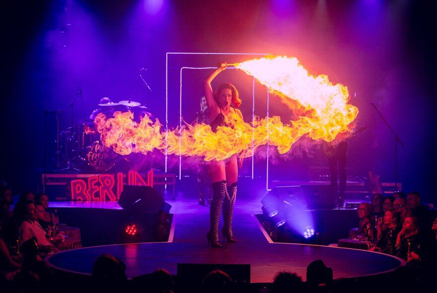 A cabaret performer twirling a fire stick on stage before a seated audience; purple lights on her.