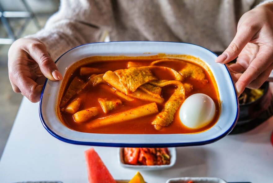 Hands holding an enamel tray with a Korean meal featuring a whole boiled egg on the side. 