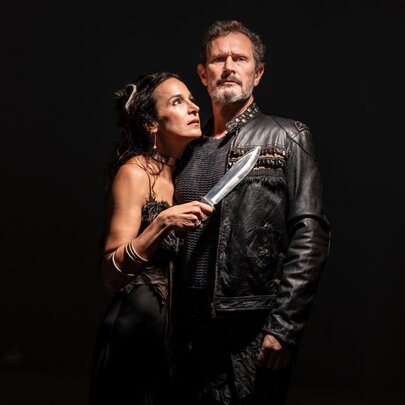 Two actors portraying characters in Shakespeare's Macbeth, with Lady Macbeth holding a knife to Macbeth's chest as she hugs him from the side. 