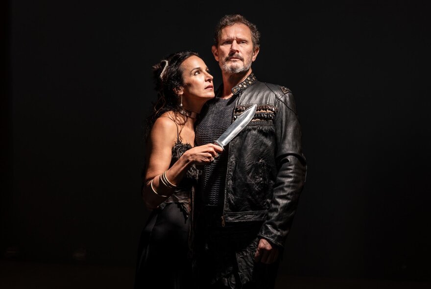Two actors portraying characters in Shakespeare's Macbeth, with Lady Macbeth holding a knife to Macbeth's chest as she hugs him from the side. 