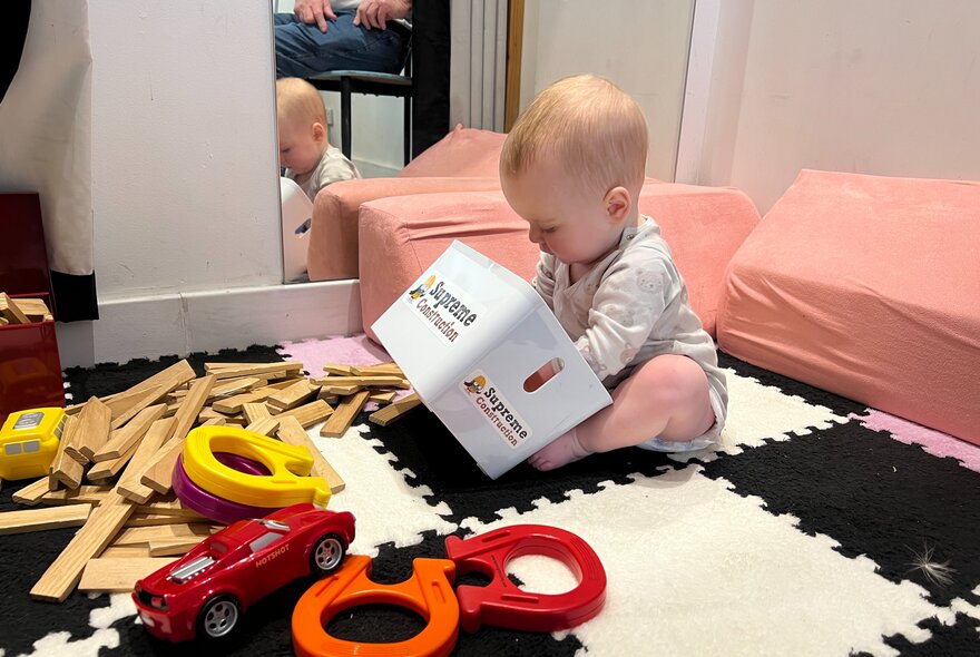 A toddler seated on a mat playing with a box and toy construction equipment.