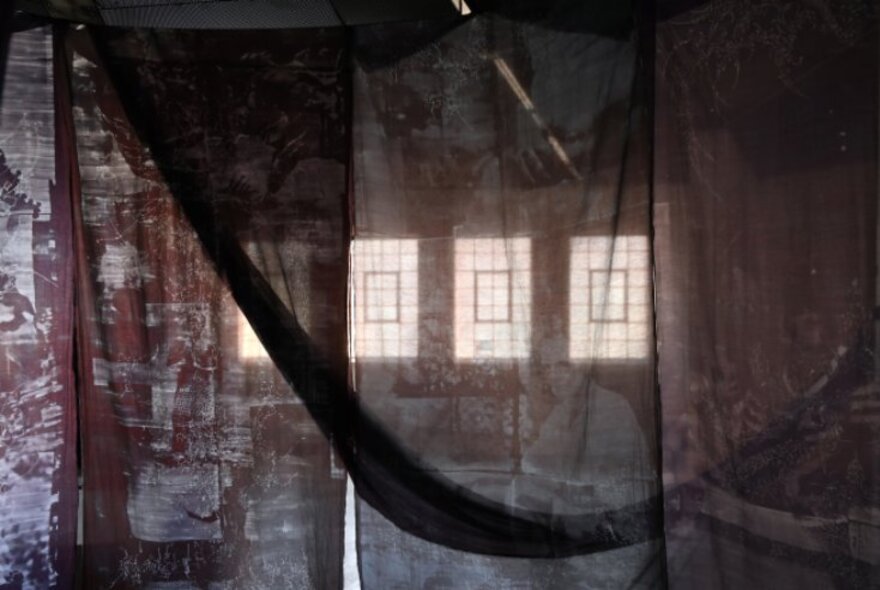 A darkened room with four windows, seen through multi-layered gauzy fabrics draped from the ceiling.