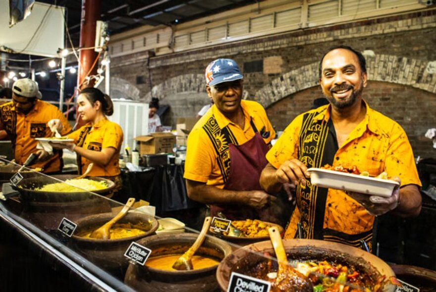 Cheerful food servers in front of large vats of curries, rice and other dishes.
