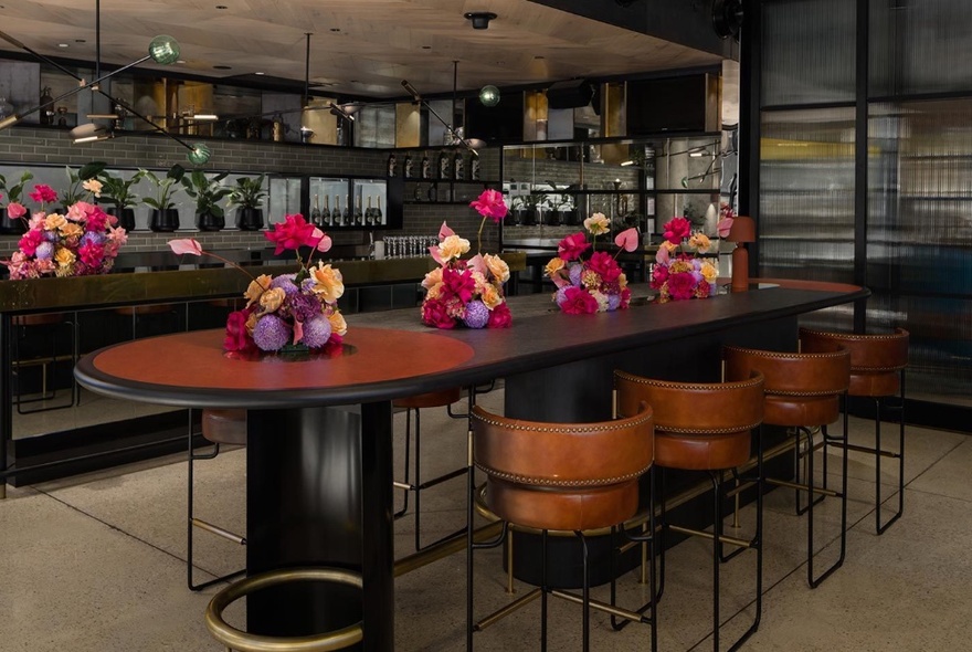 An oblong bar table with leather bar stools and pink, purple and apricot coloured flower arrangements on top.