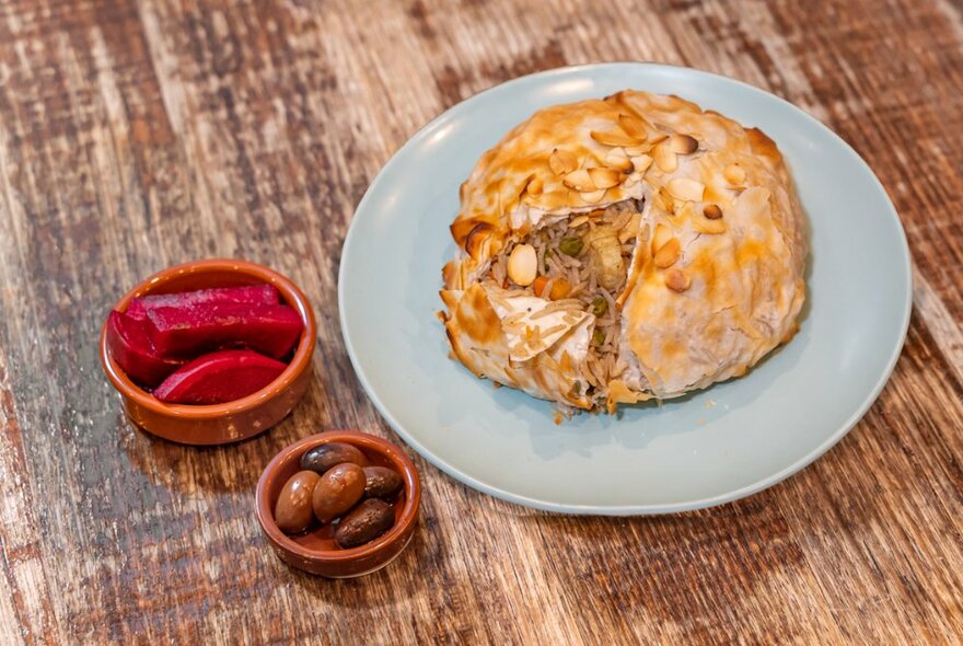 An enclosed pastry served on a plate with two small sides of pickled beets and olives, on a wooden table.