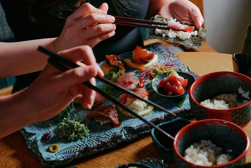 Hands holding chopsticks taking rice and items from bowls and large sushi platter.