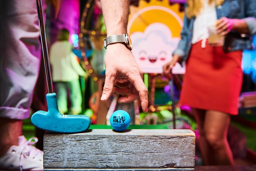 Hand placing a blue golf ball on a putting tee, with golf club nearby and people in the background.