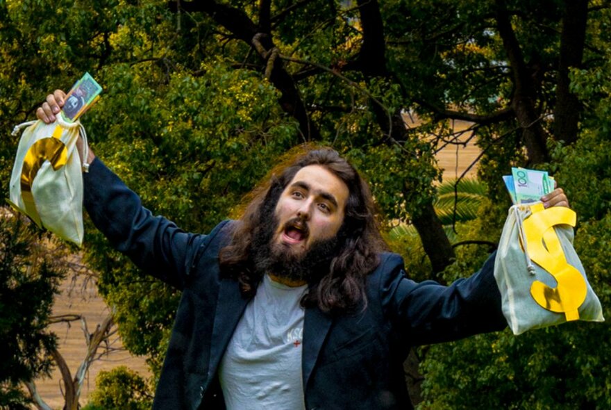 Comedian with long hair and a beard, holding cash and bags with yellow dollar signs in front of trees.