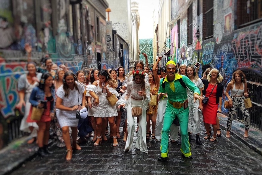 Group dancing in a laneway, led by man in bright green and yellow shiny outfit, all wearing headphones.