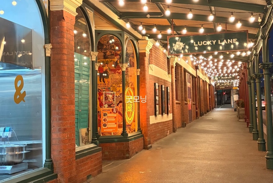An outdoor pedestrian walkway alongside a historic red brick building, illuminated by warm overhead string lights with arched shop windows.