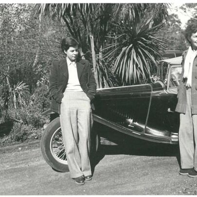 A historic black-and-white photograph shows June Barnett and another woman standing next to a vintage car around the 1950s.