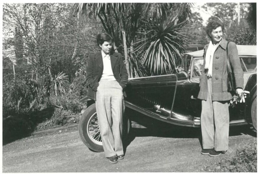 A historic black-and-white photograph shows June Barnett and another woman standing next to a vintage car around the 1950s.