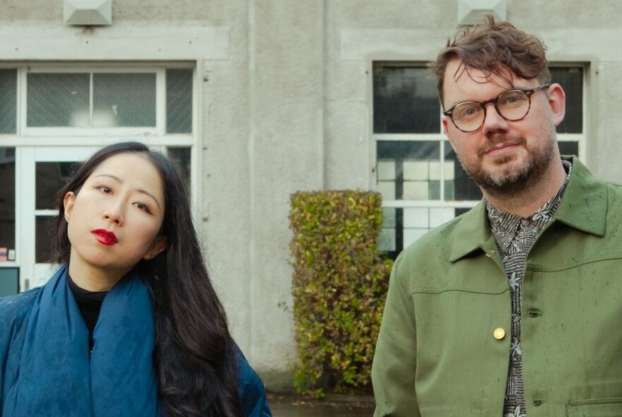 Musicians Mindy Meng Wang and Tim Shiel looking neutral in front of a building with a hedge.