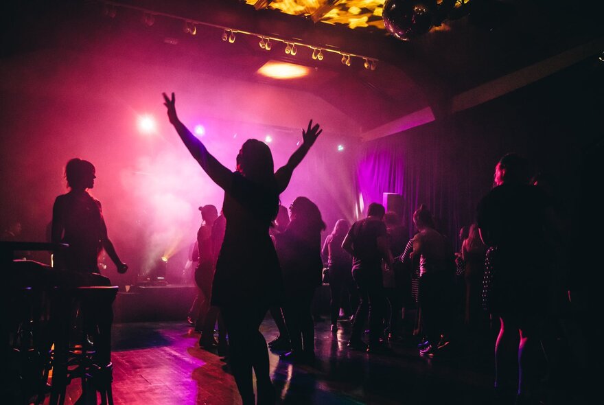 A woman with her hands in the air while people dance in front of a stage with a pink glowing light. 