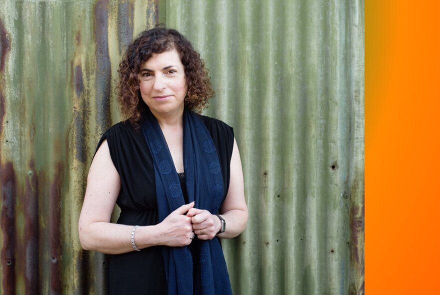 Woman wearing a black dress with shoulder length curly brown hair,smiling slightly and posed against a pale green corrugated iron fence. 