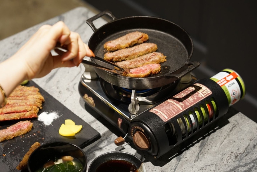 Slices of Japanese gyukatsu (grilled beef) cooking on a small tabletop gas burner, with a hand using large tweezers to turn them.