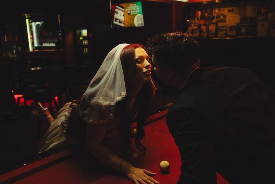 A woman wearing a white veil is leaning over a red pool table in a dimly lit bar, about to kiss a man in a dark suit who is leaning in close to her.