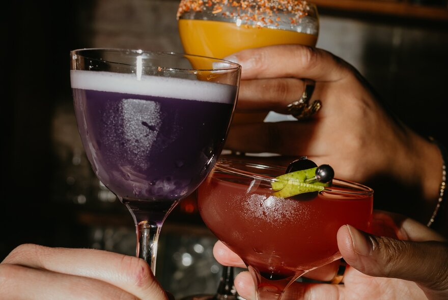 Three cocktails being bought together in a toast - one purple, one red and another orange. 