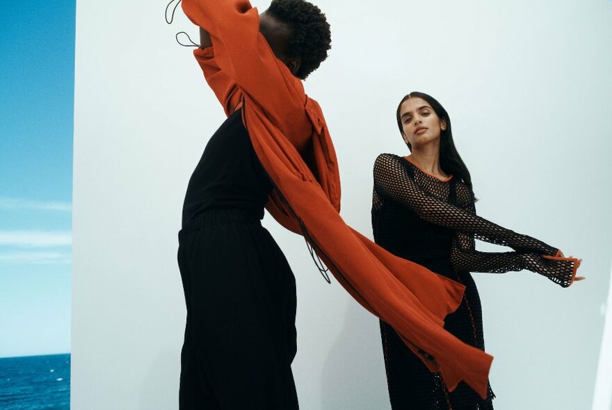 Two models wearing dark clothing, one wearing a red coat, posing against a white backdrop, with the blue sea visible in the background.