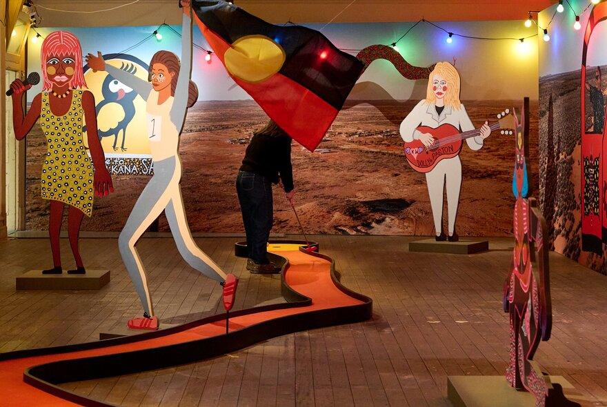 A woman playing mini golf in a room with wooden cutouts, festoon lights and an outback backdrop.
