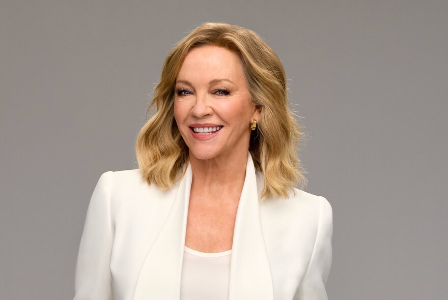 Portrait of TV personality, Rebecca Gibney, smiling, wearing a white jacket and top; grey background.