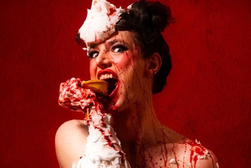 Saucy woman biting into a pie with cream and red sauce like blood dripping over her face and shoulders.