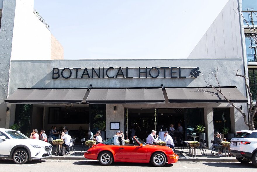 People seated at cafe tables outside Botanical Hotel in Domain Road, red sportscar parked outside.