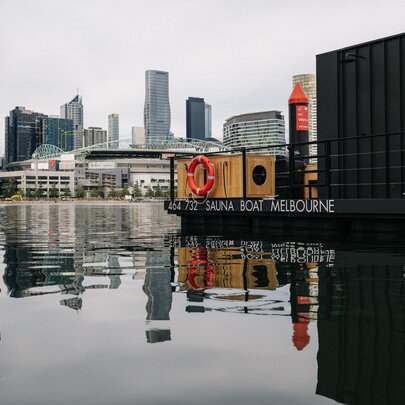 A floating sauna barge moored in Docklands harbour with city buildings in the background.