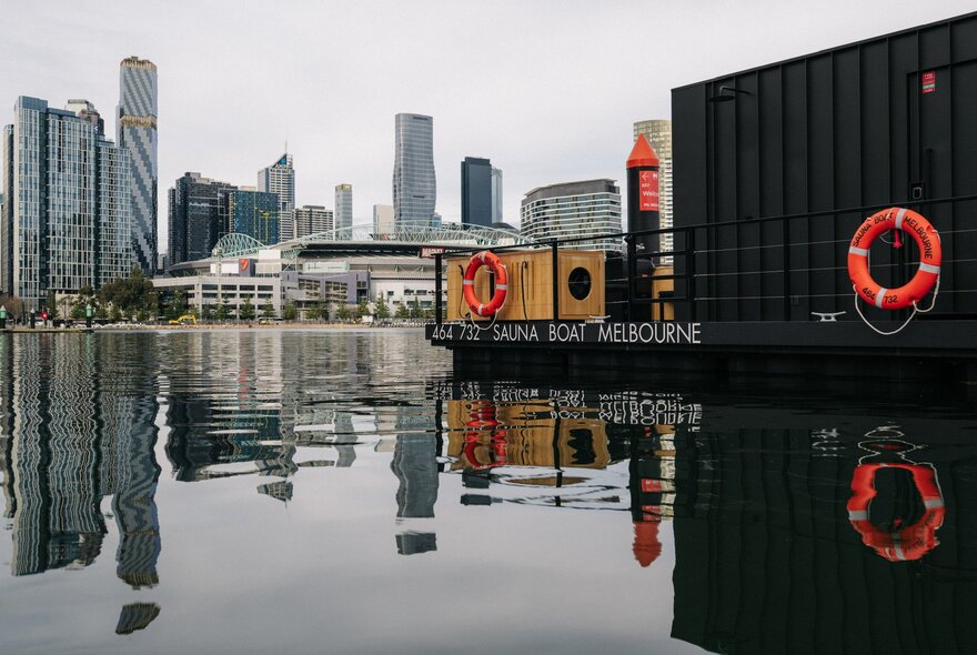 A floating sauna barge moored in Docklands harbour with city buildings in the background.