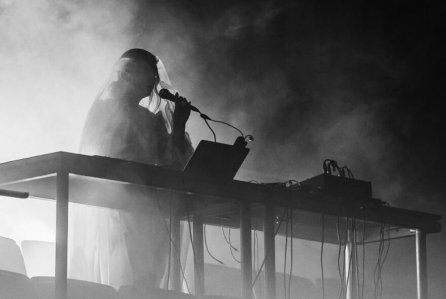 A musician silhouetted and performing on stage behind an electronic keyboard and mixing panel, a smoke effect haze surrounding them.