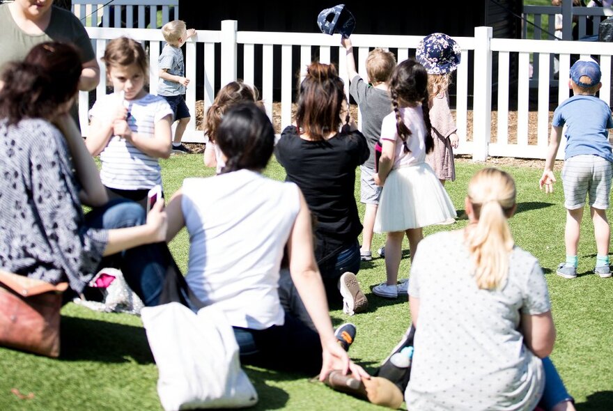 Young children and their parents sitting on the lawns at Melbourne Zoo with white picket fence and stage in the background.