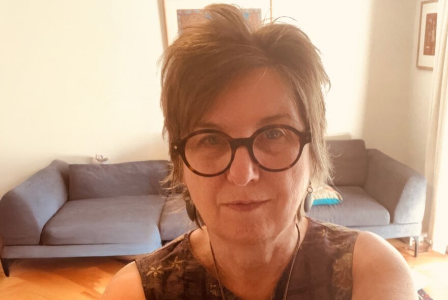 A woman with short hair and round glasses on Zoom call with her sofa in the background. 