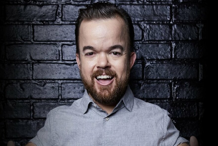 Bearded and smiling comedian Brad Williams, wearing a light grey shirt and posed against a black brick wall.