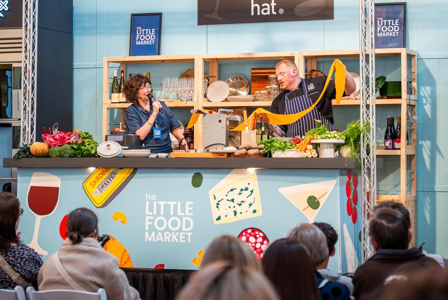 Live cooking demo with presenter speaking into a microphone while another stretches a long length of pasta.