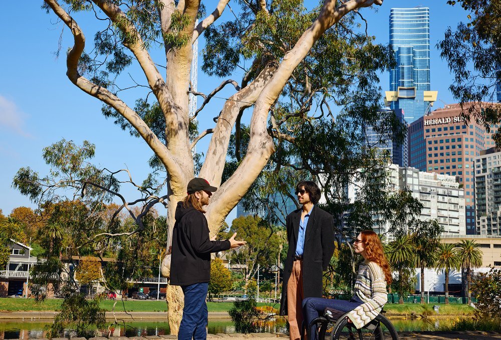 A guide talking to two people by a gumtree next to the Yarra River in the Melbourne CBD. The woman is using a wheelchair.