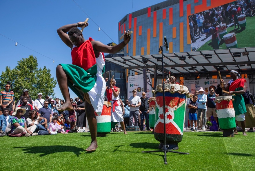 African dancers and drummers performing on a lawn with Federation Square in the background. 