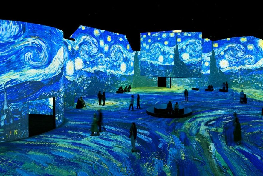 People walking around a very large space with Vincent van Gogh artwork digitally projected on all the wall and floor spaces.