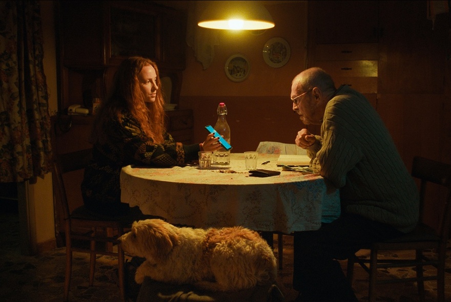 A still image from a film showing a small, dimly lit room with a woman and at a round table with a single light above them and a dog on a chair. 