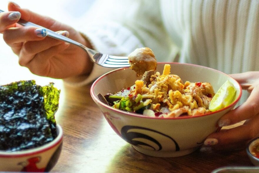 A person's hands scooping Thai food from a bowl with a fork.