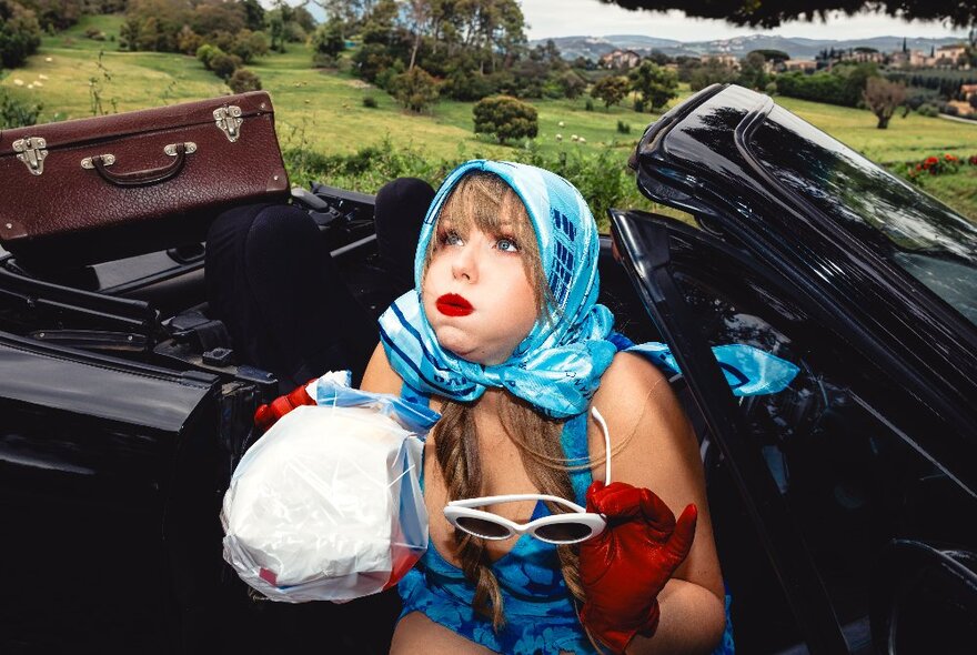 Comedian wearing headscarf, seated in an open-top car holding a paper bag and looking like she's about to vomit.