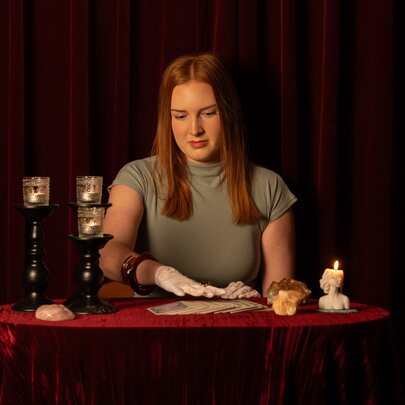 A person sitting at a small table draped in red fabric, performing a tarot card reading in front of a dark red curtain backdrop, surrounded by lit candles and crystals.