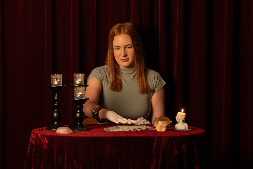 A person sitting at a small table draped in red fabric, performing a tarot card reading in front of a dark red curtain backdrop, surrounded by lit candles and crystals.