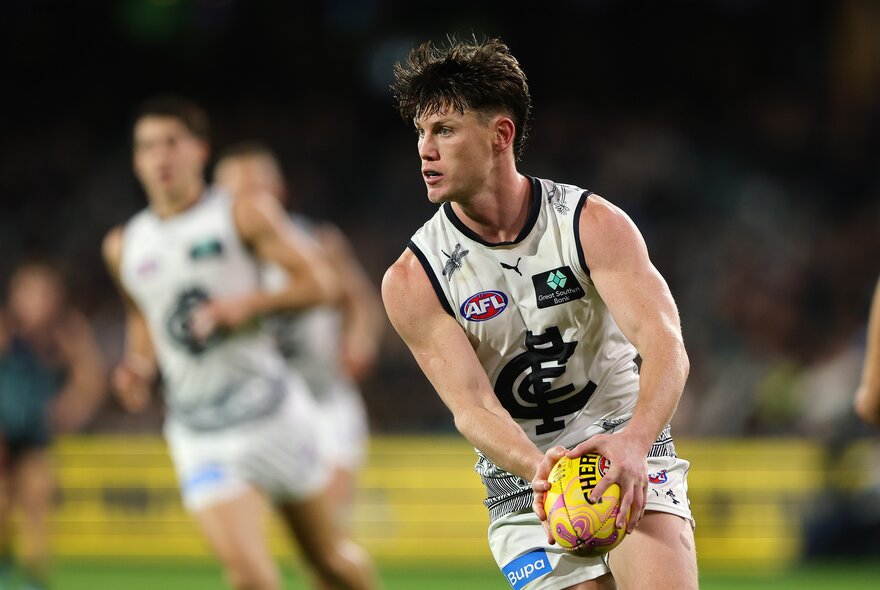 A Carlton AFL player on the field running while holding the ball, blurred players in the background.
